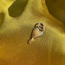 A small gold-toned metal charm or pendant with a heart-like shape and a loop at the top, placed on draped shiny gold fabric.