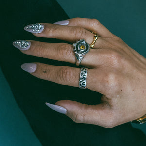 A hand model wearing a Sterling silver evil eye protection ring with faceted citrine in the eye's center, a silver leopard print stacking band, and a gold bone eternity ring.