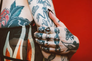 Alt model with tattoos, wearing 3 rings. A sterling silver evil eye protection ring with garnet in the eye center, a silver serpent ring with round onyx cabochon, and a thorny twig ring in silver. Red background. Sterling silver evil eye protection ring with faceted garnet in eye center, garnet ring, sterling silver ring, evil eye ring, drippy eye ring, hellhound jewelry ring, faceted garnet ring, gemstone ring, gemstone jewelry, gemstone